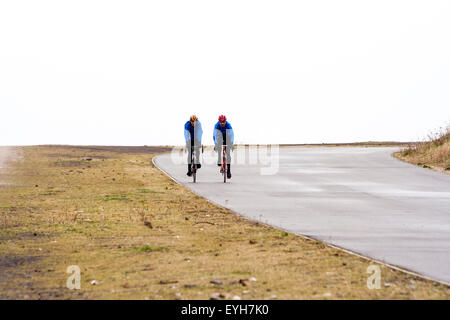 Zwei kaukasischen Radfahrer, das Tragen von Sturzhelmen, in der Nähe des Horizonts, Radfahren in Richtung Betrachter auf breiten asphaltierten unmarkierte Straße durch Grasland. Grauer Himmel. Stockfoto