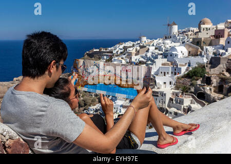Junges asiatisches Paar, das sich eine Karte auf der Terrasse des Oia Castle mit der berühmten Windmühle und dem Dorf im Hintergrund anschaut, Santorini, Griechenland Stockfoto