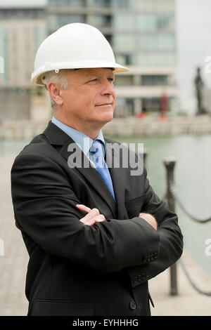 Geschäftsmann, trägt einen dunklen Anzug und weißen Schutzhelm, steht mit verschränkten Armen. Stockfoto