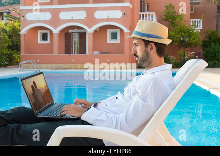 Junge Bussines Mann auf Pause während der Arbeit an seinem Laptop durch den Pool während auf Ferien tragen Stroh Hut Stockfoto