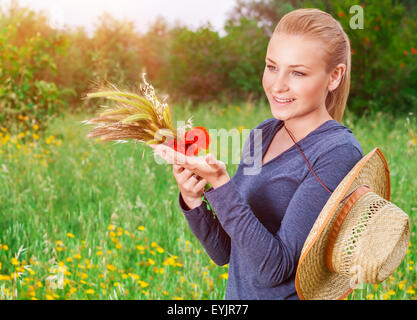 Schönen Bauernmädchen auf grünen Blumen Wiese im milden Abendlicht, reifen Ährchen des Weizens und der rote Mohn Blume in den Händen halten Stockfoto