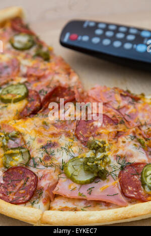 Stück Pizza mit Wurst und Speck in Pappmaché Box mit TV-Fernbedienung Stockfoto