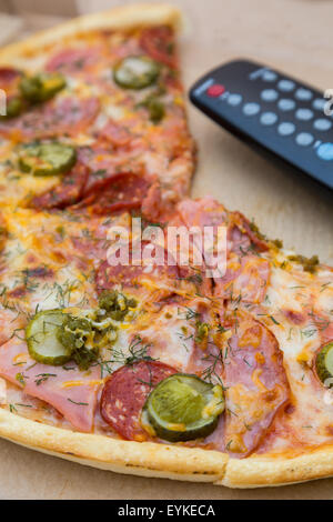 Stück Pizza mit Wurst und Speck in Pappmaché Box mit TV-Fernbedienung Stockfoto