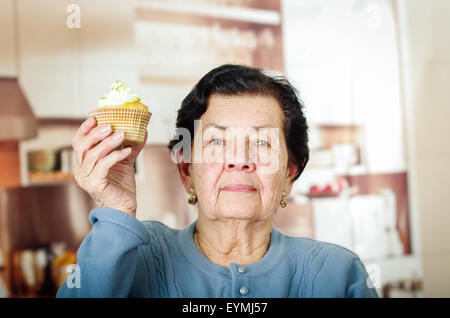 Ältere hispanic Frau mit blauen Pullover sitzen vor der Kamera hält einen gelben Cupcake mit Sahnehäubchen Stockfoto