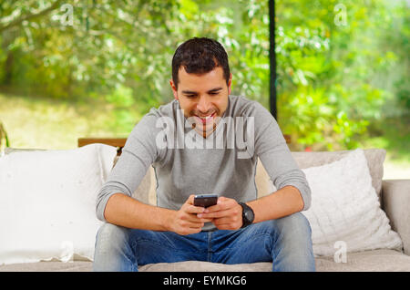 Hispanic Mann tragen Denimjeans mit grauen Pullover sitzt im Sofa gelehnt zukunftsorientiert am Handy-Bildschirm Stockfoto