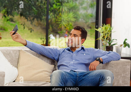 Hispanic Mann in Jeans und Hemd sitzt im Sofa für Selfie lächelnd zu Handy-Kamera zu posieren Stockfoto