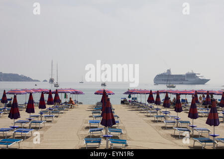 Italienischen Strand mit Reihen von leeren Sonnenliegen und das Kreuzfahrtschiff sieben Meer Mariner vor Anker Stockfoto