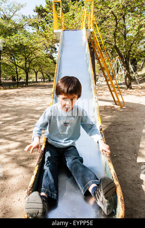 Kaukasier Kind, Junge, 7-8 Jahre alt. Vor, nach unten rutschen Spielplatz Rutsche auf seinem eigenen. Lächelnd, als er nach unten rutschen. Bäume im Hintergrund. Stockfoto