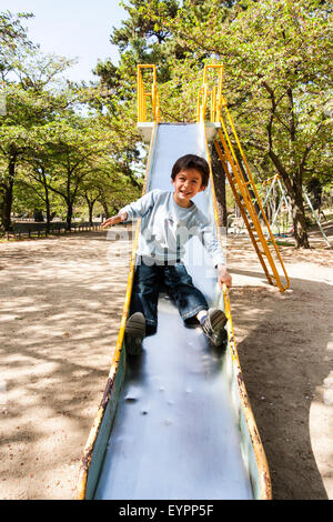 Kaukasier Kind, Junge, 7-8 Jahre alt. Vor, sie spielen - Park schieben. Augenkontakt lachte, als er allein nach unten rutschen. Bäume im Hintergrund. Stockfoto