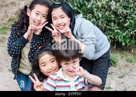 Reihe von vier lächelnden Kinder, 7-9 Jahre alt, mit Blick auf Viewer mit Blickkontakt. 3 japanische Mädchen und einen Kaukasischen jungen Alle geben Japanische Frieden Geste. Stockfoto