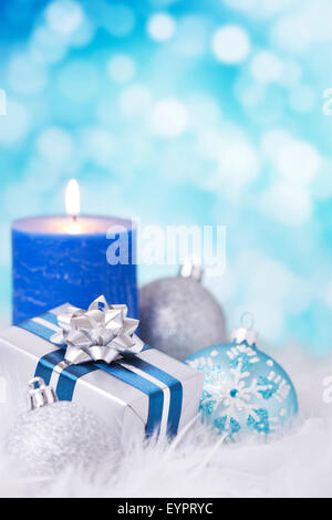 Blau und Silber Weihnachtskugeln, ein Geschenk und eine Kerze auf eine weiche federleichte Oberfläche vor defokussierten blaue und weiße Lichter. Stockfoto