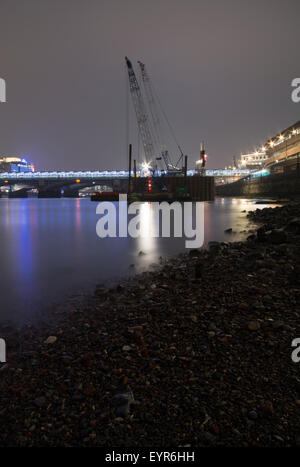 Krane und The Blackfriars Railway Bridge in der Ferne von den Ufern der Themse bei Nacht. Stockfoto