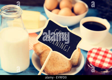 eine Tafel mit dem Text guten Morgen und einen blauen Holztisch mit einer Flasche Milch, eine Tasse Kaffee, etwas Toast in einem Teller Stockfoto