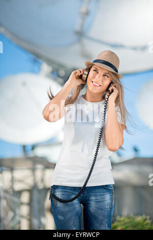 Glückliche junge Frau in Hut Musikhören im Vintage Musik Kopfhörer und tanzen vor Hintergrund der Satelliten Gerichte t Stockfoto