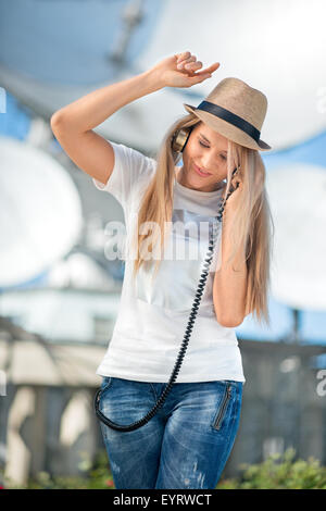 Glückliche junge Frau in Hut Musikhören im Vintage Musik Kopfhörer und tanzen vor Hintergrund der Satelliten Gerichte t Stockfoto