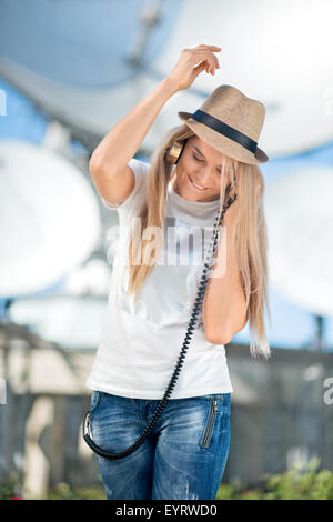 Glückliche junge Frau in Hut Musikhören im Vintage Musik Kopfhörer und tanzen vor Hintergrund der Satelliten Gerichte t Stockfoto