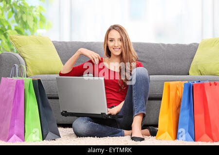 Junge Frau hält einen Laptop und sitzt vor einem Sofa mit einem Bündel von Einkaufstüten herum zu Hause Stockfoto