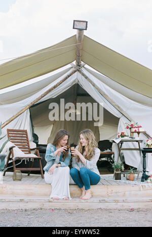 Zwei lächelnde Frauen draußen ein großes Zelt, lachen und ein Glas Wein zu sitzen. Stockfoto