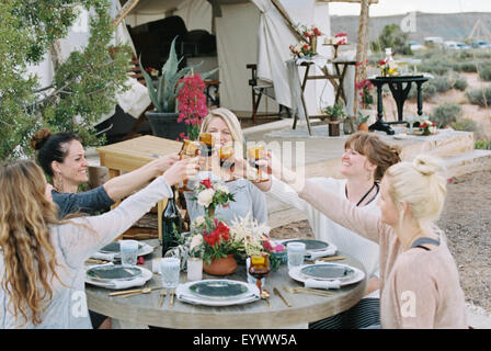 Eine Gruppe von Frauen genießen eine Mahlzeit im Freien durch ein großes Zelt in einer Wüstenlandschaft, einen Toast durch klirrende Gläser. Stockfoto