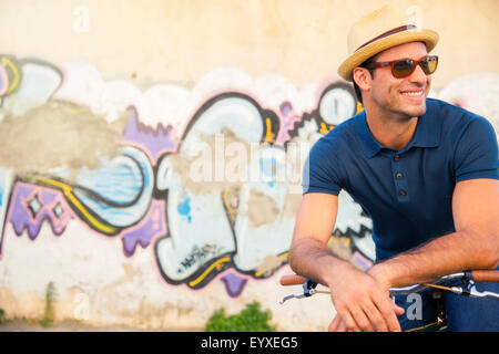Lächelnder Mann in Hut und Sonnenbrille stützte sich auf dem Fahrrad neben urban Graffitiwand Stockfoto