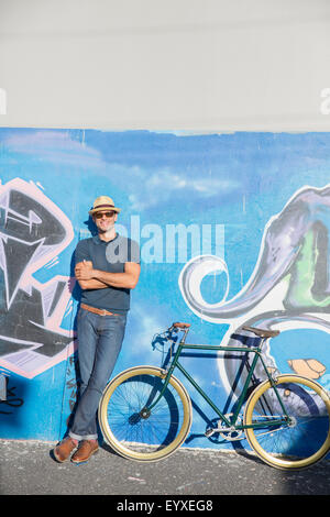Porträt, Lächeln Mann in Hut und eine Sonnenbrille stützte sich auf urban Graffitiwand neben dem Fahrrad