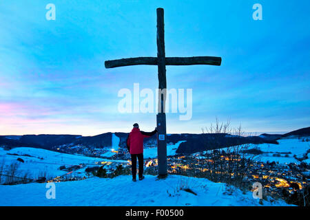 Mann neben Kreuz auf Oerenberg Berggipfel und genießen Sie die Aussicht auf Winterlandschaft am Abend, Willingen, Sauerland, Hessen, Deutschland Stockfoto