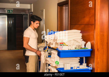 Junger Mann schob einen Haushalt Wagen beladen mit saubere Handtücher, Wäsche und Reinigungsgeräte in einem Hotel, als er das Zimmer services Stockfoto