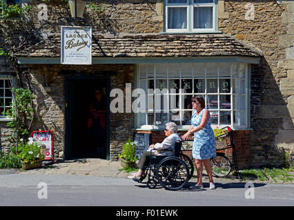 Alte Frau im Rollstuhl vorbei die Lacock-Bäckerei im Dorf Lacock, Wiltshire, England UK geschoben Stockfoto