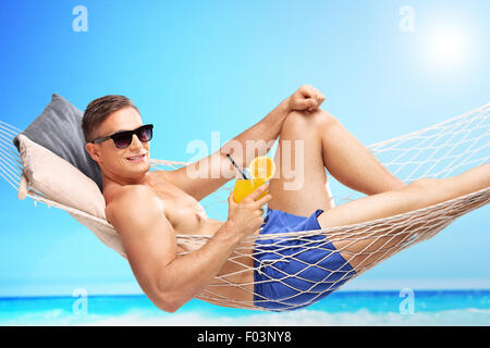 Hübscher junger Mann hält einen orangenen Cocktail und liegen in der Hängematte an einem sonnigen Strand durch einen Ozean Stockfoto
