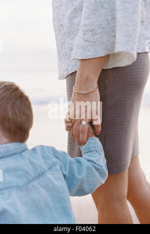 Frau stehend auf einem Sandstrand am Meer, ihren kleinen Sohn Hand hält. Stockfoto