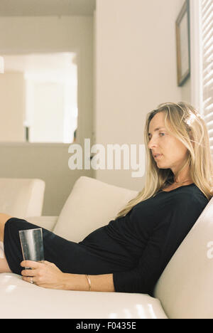 Frau mit langen blonden Haaren trug ein schwarzes Kleid, sitzt auf einem Sofa mit einem Glas. Stockfoto