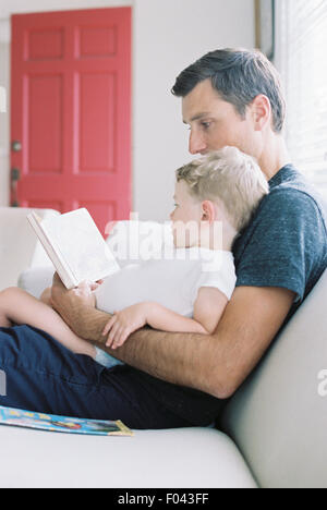 Mann sitzt auf einem Sofa mit seinem Sohn sitzt auf seinem Schoß, hält ein Buch, eine Geschichte zu lesen. Stockfoto