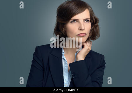 Attraktive nachdenkliche Frau sitzt mit ihr Kinn auf ihre Hand starrte in den Weltraum mit einem ernsten Ausdruck über grau Stockfoto