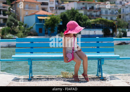 Kleines Mädchen mit rosa Hut sitzt auf einer blauen Bank am Meer Stockfoto