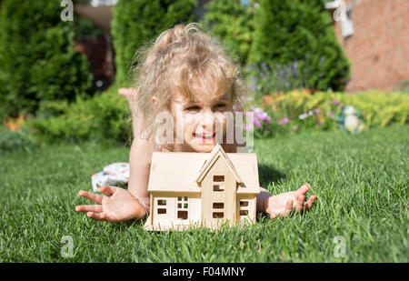 Porträt von kleinen Mädchen liegend auf dem Rasen mit Holzmodell des Hauses in Händen, Sommer im Freien, neues Wohnkonzept Stockfoto