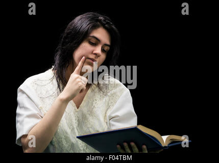 Latina Teen im weißen Kleid hält ein blaues Buch mit Gold Rand, halten einen Finger auf ihre Wange und stand vor einem schwarzen Stockfoto