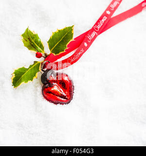 herzförmige Rote Christbaumkugel mit Holly und Frohe Weihnachten Ribbon auf Schnee Stockfoto
