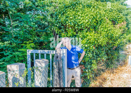 Kaukasischer Mann mittleren Alters, elegant und sportlich kleiden einen blauen Polo hat einen nachdenklichen Blick während Kopf und lehnte sich gegen ein Stahltor in der italienischen Landschaft im Sommer Stockfoto