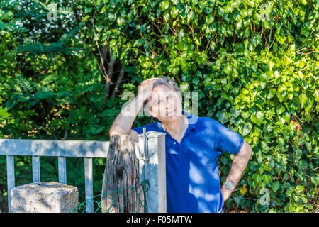 Kaukasischer Mann mittleren Alters, elegant und sportlich kleiden einen blauen Polo hat einen nachdenklichen Blick während Kopf und lehnte sich gegen ein Stahltor in der italienischen Landschaft im Sommer Stockfoto