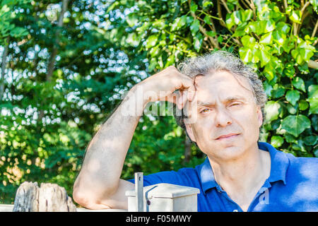 Kaukasischer Mann mittleren Alters, elegant und sportlich kleiden einen blauen Polo hat einen nachdenklichen Blick während Kopf und lehnte sich gegen ein Stahltor in der italienischen Landschaft im Sommer Stockfoto