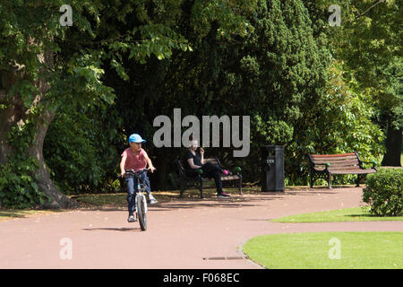 Ein kleiner Junge, der sein Fahrrad durch die West-Park mit einer Dame, beobachtete ihn von einer Bank in Wolverhampton West Midlands UK Stockfoto