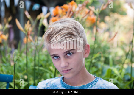 Ein gut aussehender blonder Junge schaut in die Kamera in einem Englsih Landschaftsgarten mit Pflanzen und Girlanden im Hintergrund Stockfoto