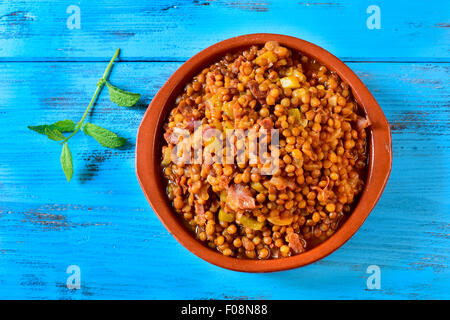 High-Angle Shot eine Steingut-Schale mit spanischen Linsen-Eintopf mit Serrano-Schinken auf einem blauen rustikalen Holztisch Stockfoto