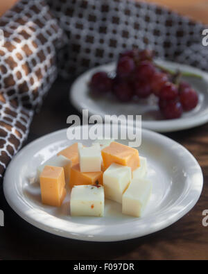 Ein weißen Teller mit verschiedenen gewürfelten Käse sitzt auf einem braunen hölzernen Tischplatte.  Ein weißer Teller rote kernlose Trauben am Rebstock ist Stockfoto