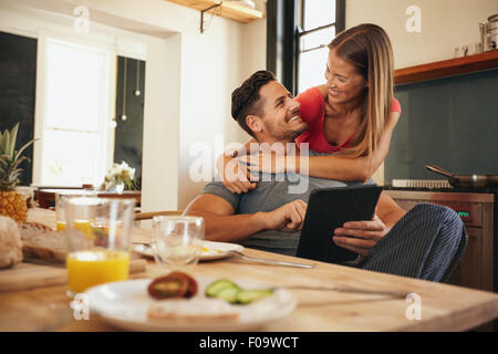Schuss des Liebens junges Paar in der Küche mit Frühstückstisch Morgen. Mann mit digitalen Tisch, während Frau umarmt ihn vom Wald Stockfoto