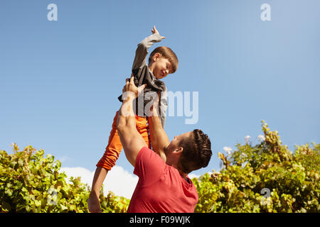 Aufnahme des jungen Mann, der seinen kleinen Sohn hoch in Luft gegen Himmel an einem sonnigen Tag hält. Glücklicher Vater und Sohn Spaß im freien Stockfoto