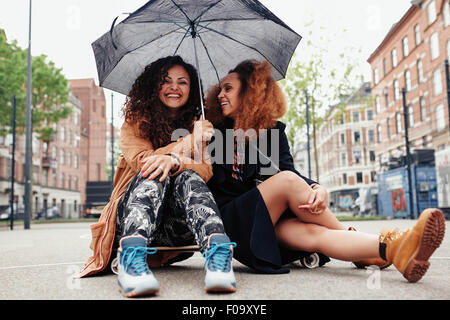 Zwei Freundinnen Zusammensitzen auf Skateboard. Lächelnde junge Frauen im Freien auf Stadtstraße mit Regenschirm. Stockfoto