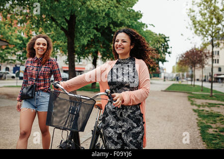 Lächelnde junge Freunde zu Fuß auf der Stadtstraße, treibt eine Frau ihr Fahrrad. Zwei junge Mädchen der Hipster mit Zyklus mit f Stockfoto