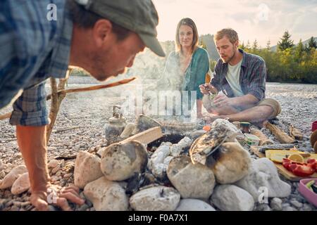 Erwachsene sitzen um Lagerfeuer zubereiten und bläst auf Glut Stockfoto