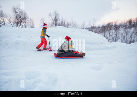 Mutter mit zwei Söhnen auf Schlitten und Schnee Scooter auf schneebedeckten Hügel, Hemavan, Schweden Stockfoto